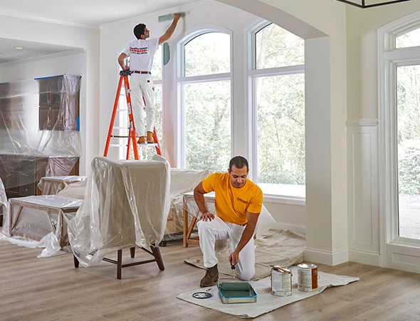 One worker mixing paint while another applies paint on wall using roller while standing on stairs