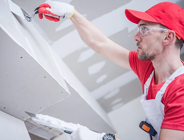A worker in protective glasses and a red cap applies plaster to a ceiling joint using a trowel. He wears a red shirt and white overalls, focused on his task.