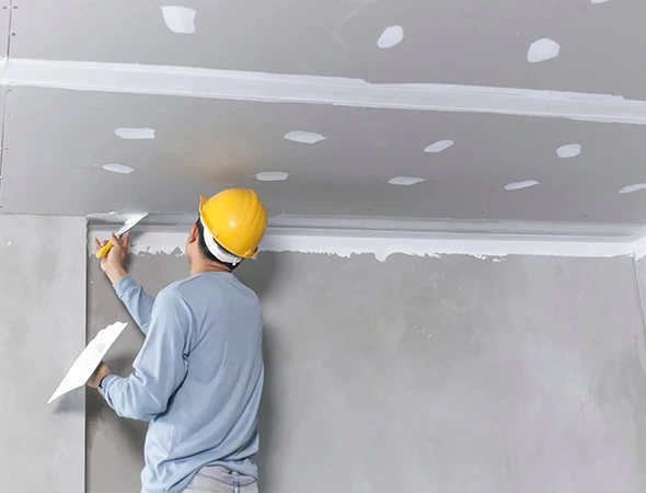 A worker in a yellow hard hat uses a trowel to smooth drywall joint compound on a ceiling corner. The scene conveys precision and focus in construction.