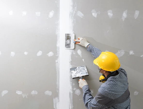 Worker in a yellow hard hat applies plaster with trowels to a seam on a drywall surface, suggesting construction or renovation work in progress.