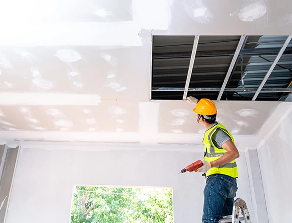 A construction worker in a yellow vest and hard hat stands on a ladder in a room, repairing a ceiling with tools. Sunlight streams through a window.