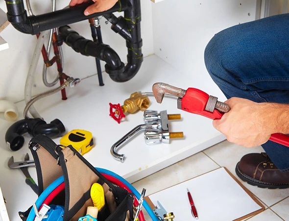 A plumber kneels on the floor, fixing pipes under a kitchen sink with a red wrench. Nearby are tools, fittings, and a clipboard, conveying a busy work scene.