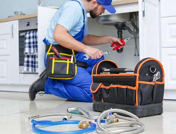 A plumber in blue overalls kneels by a kitchen sink, using a wrench. A tool bag and hoses are spread out on the floor, indicating active repair work.