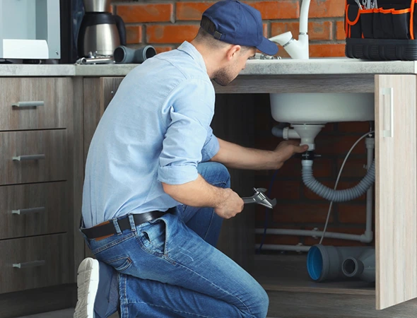 A plumber in a blue cap and shirt kneels under a kitchen sink with tools, focused on fixing a pipe. The setting is casual and practical, conveying diligence.