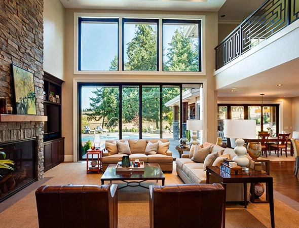Two-story living room with large windows showcasing a lush green view. Cozy beige sofas and chairs surround a coffee table. A stone fireplace and open dining area create a warm ambiance.