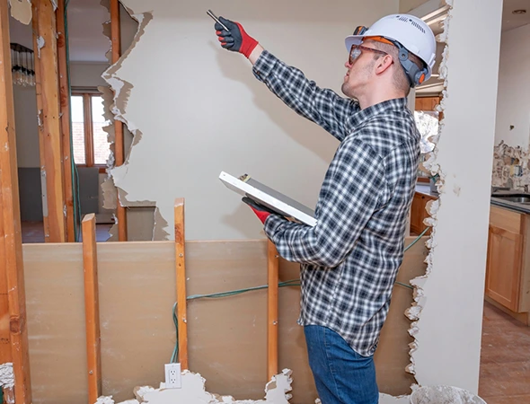 A construction worker in Rock Hill NC wearing a hard hat and glasses examines a partially demolished wall while holding a clipboard, suggesting an inspection or renovation.